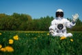 Futuristic astronaut in a helmet sits on a green lawn among flowers. Greetings to the camera