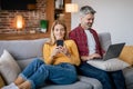 Gadget addiction at home. Smiling busy adult european man with laptop, lady with phone chatting