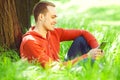 Gadget freak concept. Portrait of smiling young man in casual clothing reading e-book, sitting in green grass under tree in the Royalty Free Stock Photo