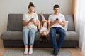 Gadgets addiction. Young family of three holding and using different electronic devices while sitting on sofa. Parents and their