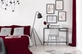 Gallery of illustrations on a white wall above a small desk which is next to a black metal lamp and a burgundy bed in a modern b