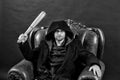 Gangster guy strike with bat weapon. Bearded man with baseball bat sit in armchair. Criminal wear hood in coat. Crime or