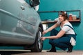 Garage work. Young smiling woman mechanic in blue coveralls squatting and repair tire with ratchet wrench. The Royalty Free Stock Photo