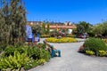 A garden with a tall metal green red and yellow rooster and a giant wooden chair with colorful flowers, lush green trees