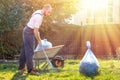 The gardener in the blue uniform is cleaning the yard.On the grass is a cart with compost and a package of garbage. Bright