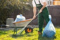 Gardener in green form cleans leaves in the yard. Beside him is a cart with compost