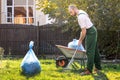 The gardener in the green uniform is cleaning the yard.On the grass is a cart with compost and a package of garbage