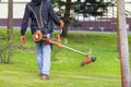 A gardener with a trimmer on a shoulder suspension walks through the garden between the trees and mows the growing grass on a