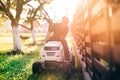 Gardner riding lawnmower and cutting grass during sunset golden hour. Details of gardening with sunrays