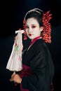 Geisha in black kimono with red flowers in hair holding traditional asian umbrella isolated on black Royalty Free Stock Photo