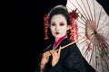 Geisha in black kimono with red flowers in hair holding traditional asian umbrella isolated on black with copy space Royalty Free Stock Photo