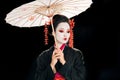 Geisha in black kimono with red flowers in hair holding traditional asian umbrella and looking down isolated on black Royalty Free Stock Photo