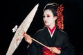 Geisha in black kimono with red flowers in hair looking at traditional asian umbrella isolated on black Royalty Free Stock Photo