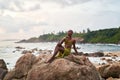 Gender fluid black person poses gracefully sitting on high rock above ocean at sunset. Androgynous ethnic fashion model