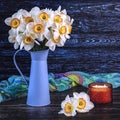 A bouquet of daffodil flowers in a blue vase, a colorful shawl, a yellow candle on a dark wooden background.Generated image