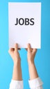 Woman holding sheet of paper with word JOBS on blue background, closeup. Career Royalty Free Stock Photo