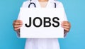 Woman holding sheet of paper with word JOBS on blue background, closeup. Career Royalty Free Stock Photo