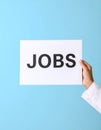 Woman holding sheet of paper with word JOBS on blue background, closeup. Career Royalty Free Stock Photo