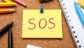 Note with word SOS and stationery on cork board, closeup Royalty Free Stock Photo