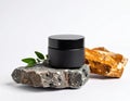 Black cylindrical cosmetic jar on a rough, textured gray stone, accompanied by green leaves. Royalty Free Stock Photo
