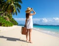 A woman in a white dress and straw hat walks along a pristine beach with a rolling suitcase. Royalty Free Stock Photo