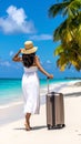 A woman in a white dress and straw hat walks along a pristine beach with a rolling suitcase. Royalty Free Stock Photo