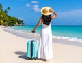 A woman in a white dress and straw hat walks along a pristine beach with a rolling suitcase. Royalty Free Stock Photo