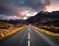 A straight, empty road leads towards a range of rugged, snow-capped mountains under a dramatic, cloud-filled sky Royalty Free Stock Photo