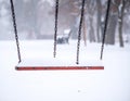 Red swing covered in snow hangs from chains, set in a snowy park Royalty Free Stock Photo