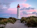 Tall white lighthouse with a red lantern room, standing amidst grassy sand dunes at sunset Royalty Free Stock Photo