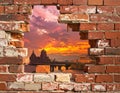 A section of a brick wall contains a large hole offering a view of a historic cityscape at sunset Royalty Free Stock Photo
