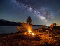 A nighttime scene by a lakeside featuring a campfire surrounded by two empty chairs. Royalty Free Stock Photo