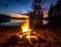 Campfire beside a tranquil lake at sunset, surrounded by tall silhouetted pine trees Royalty Free Stock Photo