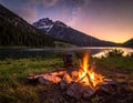 A tranquil lakeside setting features a glowing campfire surrounded by rocks, with a camping chair nearby Royalty Free Stock Photo