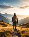 A man with a yellow backpack and hiking poles walks along a mountain trail, surrounded by grassy hills and alpine peaks Royalty Free Stock Photo