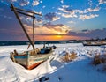 An old, weathered wooden boat rests on a snowy beach, facing a vibrant sunset over a calm sea Royalty Free Stock Photo