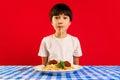 Korean boy, child in white t-shirt sitting at table and eating spaghetti with meatballs against red background. Enjoying