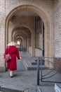 Lone woman in burgundy coat with cup of coffee walking briskly through covered arched enfilade