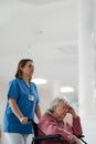 Nurse pushing senior patient in wheelchair across hospital corridor. Emotional support for elderly woman. Royalty Free Stock Photo