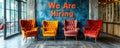 Four modern chairs against a grey wall with the words We Are Hiring written on it, signifying job opportunities and