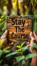 Motivational Stay The Course sign held up with two hands, promoting persistence and dedication, set against a lush green