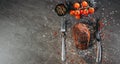 A generous piece of grilled steak on a table with fork and knife cutlery and green and red hot peppers, a sprig of cherry tomatoes