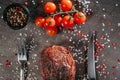 Generous piece of grilled steak on a table with fork and knife cutlery and green and red hot peppers, a sprig of cherry tomatoes,