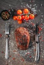 Generous piece of grilled steak on a table with fork and knife cutlery and green and red hot peppers, a sprig of cherry tomatoes,
