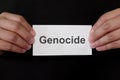 Genocide mug shot concept. Criminal hands holding paper placard with written word in dark black background.