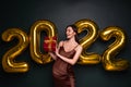 Georgeous brunette lady in beautiful dress holding gift box on black studio background with golden 2022 balloons