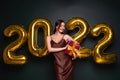 Georgeous brunette lady in beautiful dress holding gift box on black studio background with golden 2022 balloons