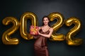 Georgeous brunette lady in beautiful dress holding gift box on black studio background with golden 2022 balloons