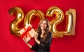 Georgeous millennial lady in beautiful dress holding gift box on red studio background with golden 2021 balloons