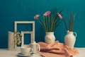 Gerbera, dry daffodils, white photo frame, a cup with coffee and a teapot and two vases with flowers on a blue background Royalty Free Stock Photo